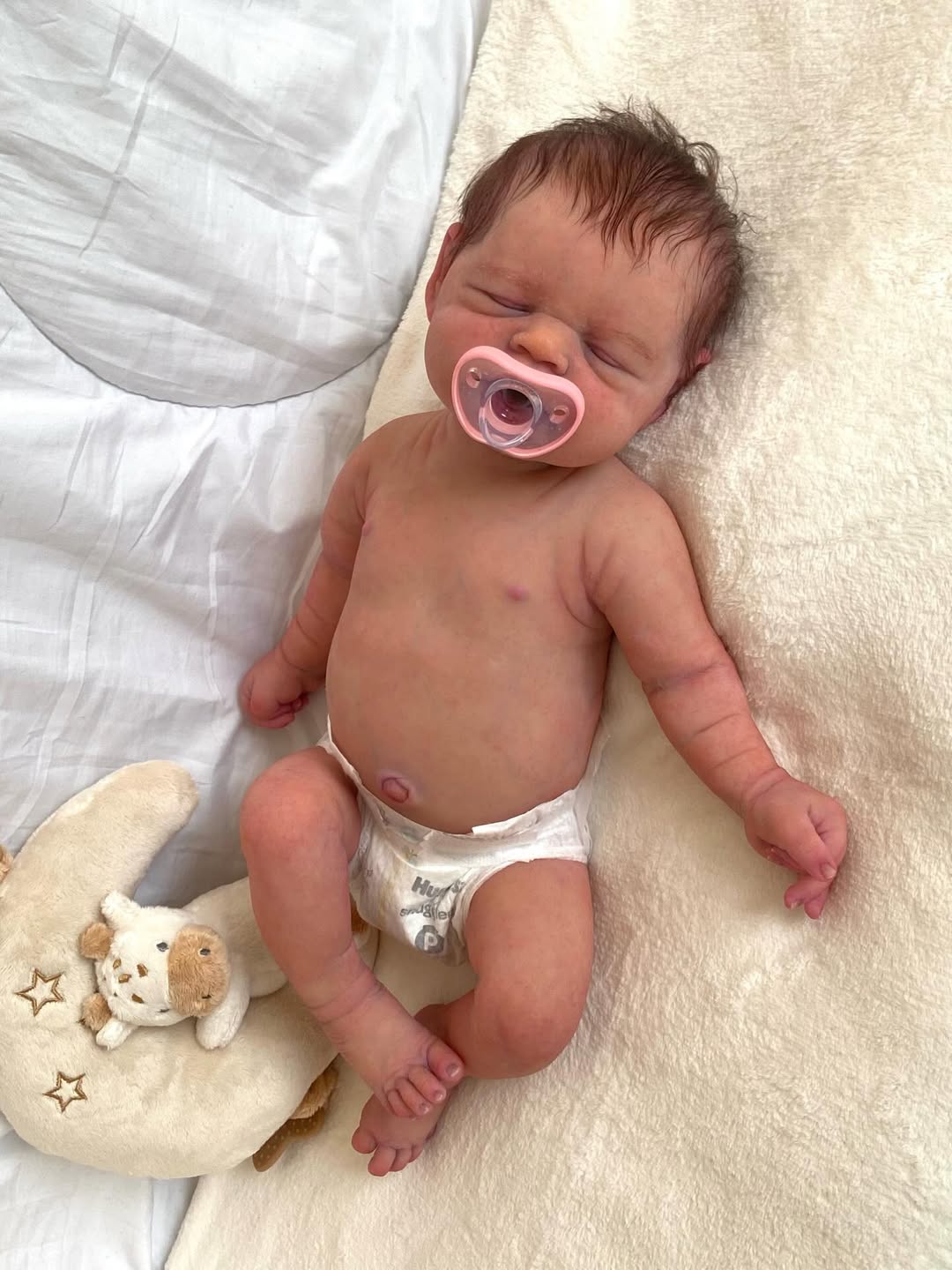 Baby Leo, a limited edition 18.3-inch full body silicone reborn boy doll with hand-rooted dark mohair, sleeping peacefully on a soft cream blanket in a white diaper while using a pink pacifier by Reborn Heaven.