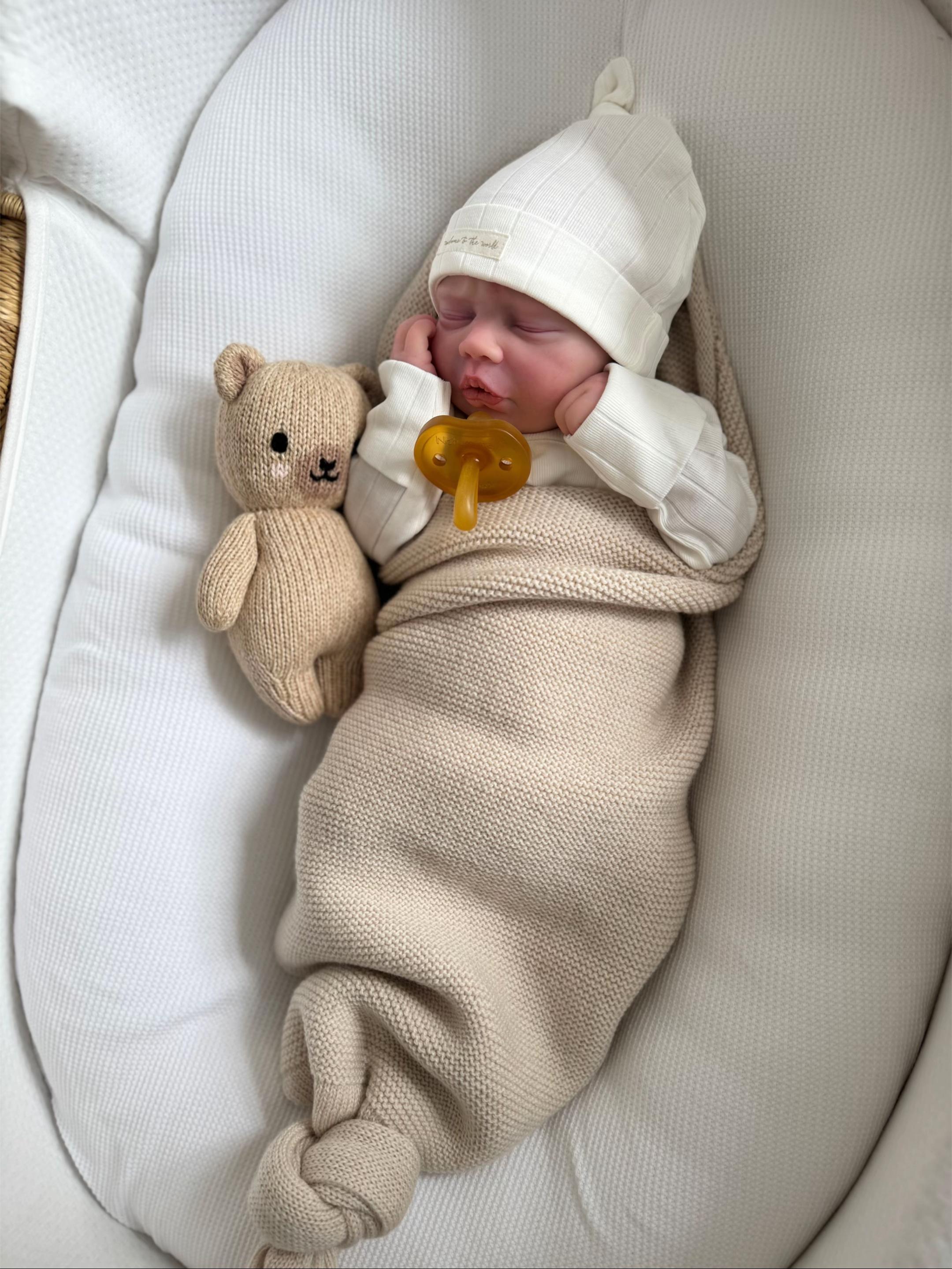Baby Ernie, an in-stock 17-inch realistic full body silicone drink and wet system baby doll with hand painted hair sleeping in a beige swaddle blanket with a white knit hat, high-end lifelike companion toy by Reborn Heaven.