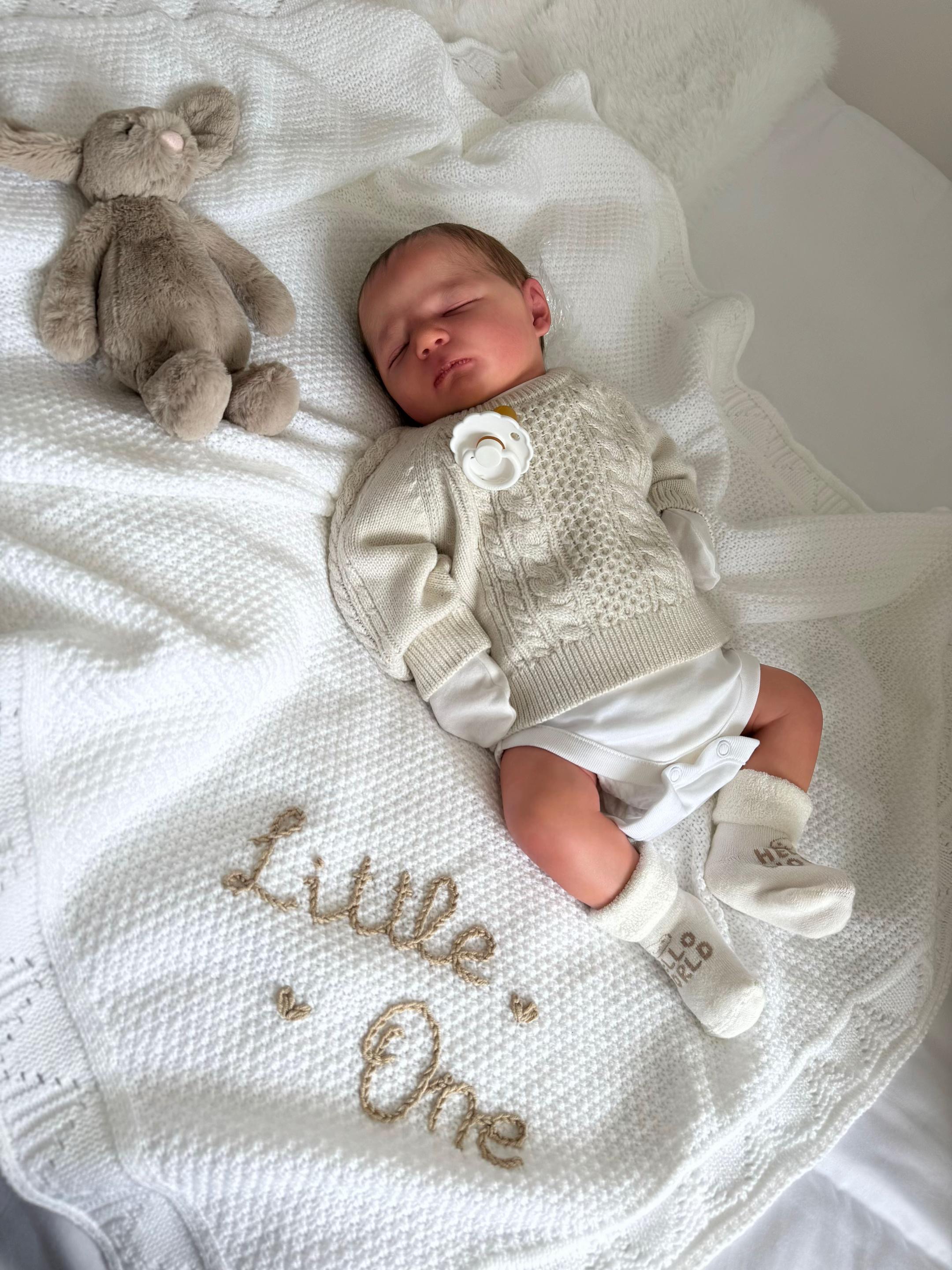 Baby Theo, an in-stock 17-inch realistic full body vinyl baby boy reborn doll with hand rooted hair sleeping soundly on a white knit blanket next to a plush toy by Reborn Heaven.