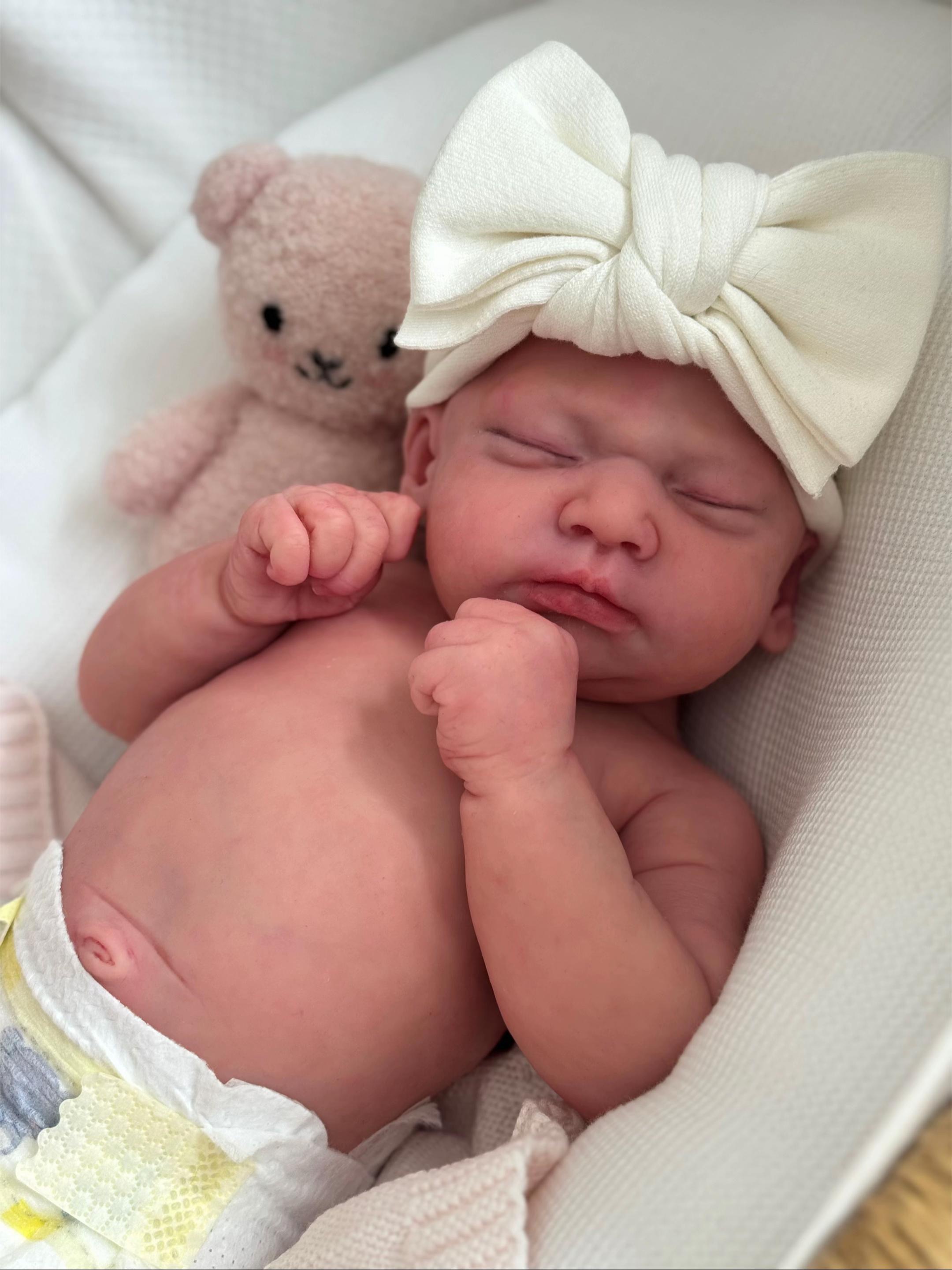 Baby Aria, an in-stock 17.1-inch realistic full body silicone baby girl reborn doll featuring a bald design and sleeping peacefully in a soft white lounge nest wearing a luxury cream bow and diaper by Reborn Heaven.