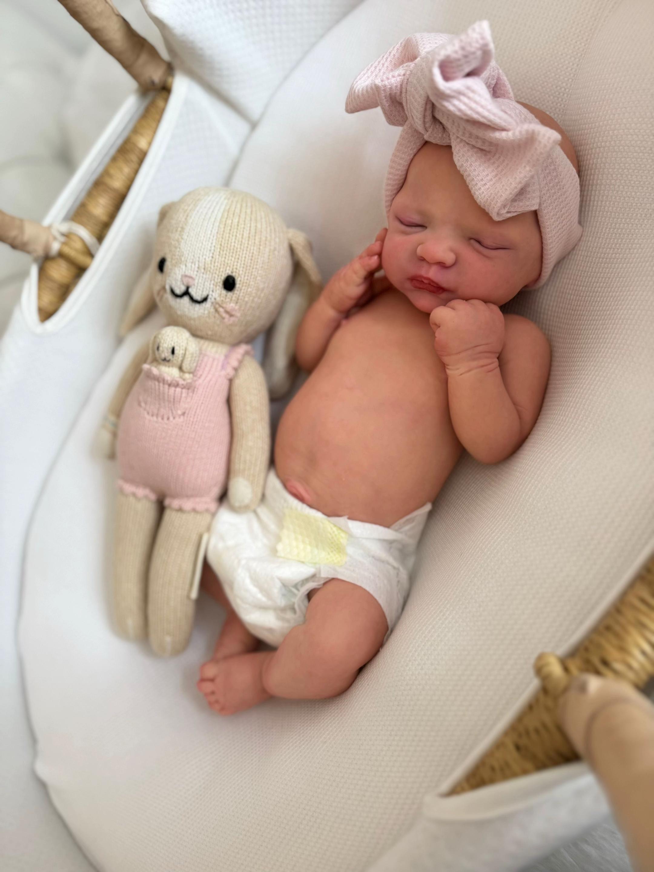 Baby Lola, an 18-inch full body silicone baby girl reborn doll with a bald head, sleeping in a white bassinet in a diaper and pink bow next to a knit bunny toy by Reborn Heaven.